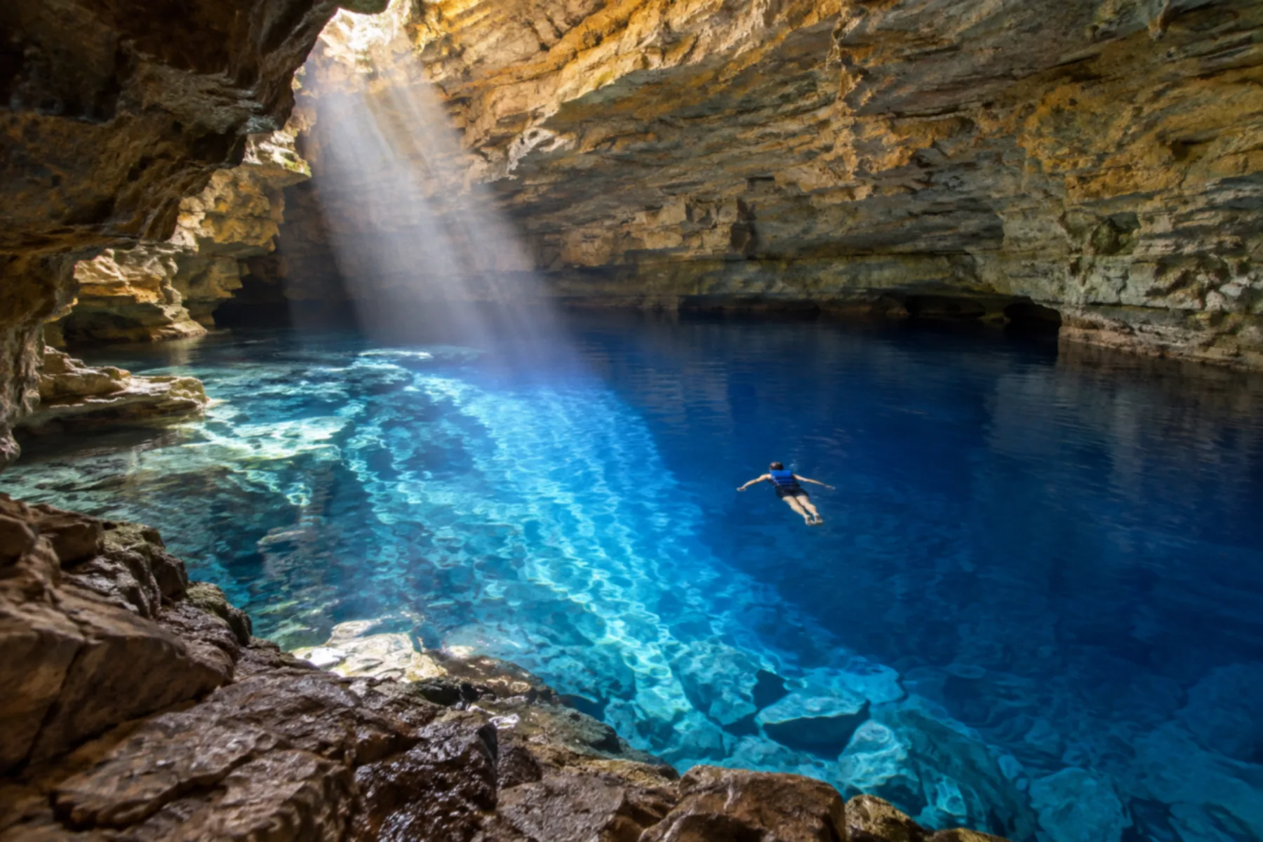 Interior de caverna com feixe de luz atravessando a água azul-cobalto e uma pessoa flutuando de colete sobre o poço.