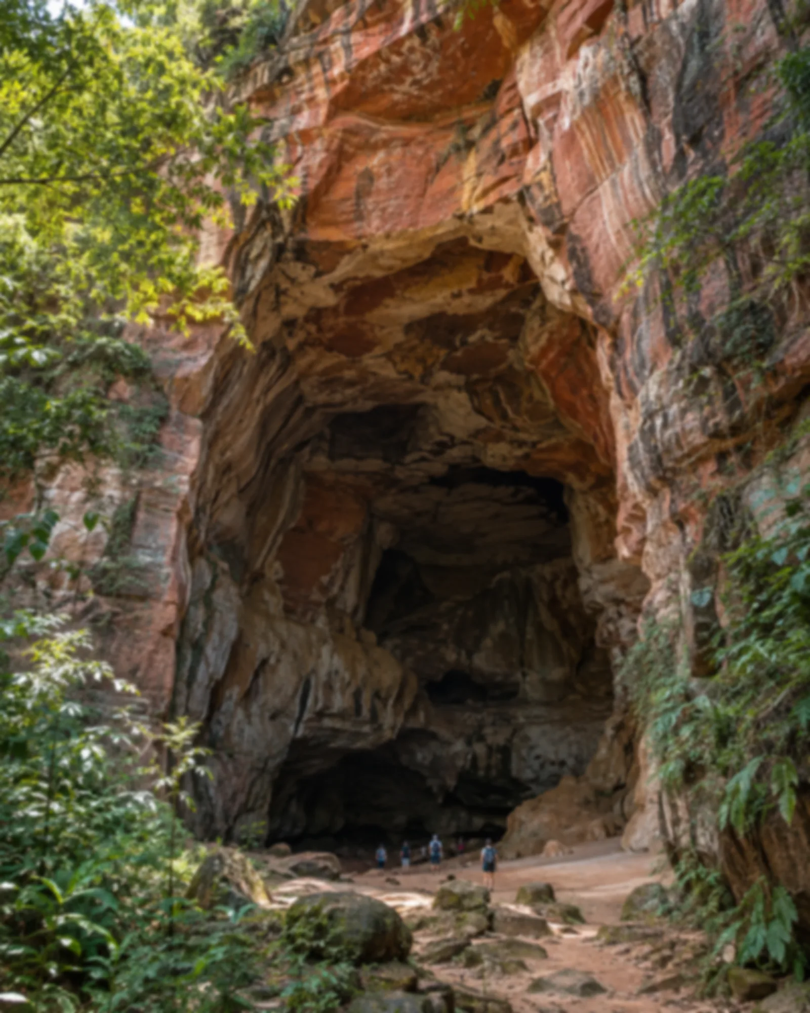 Entrada monumental de caverna de arenito avermelhado com vegetação ao redor e visitantes pequenos na boca da gruta.